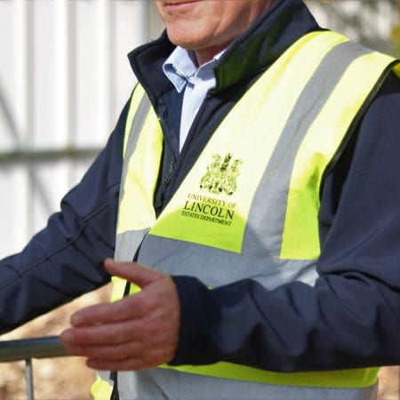 Image of a person gesturing at something. They are wearing a high visibility reflective vest with the university logo on it