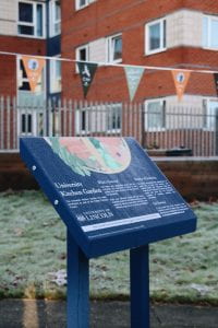 Image of a blue lectern style sign in a green frosty area. There are planters in the background.